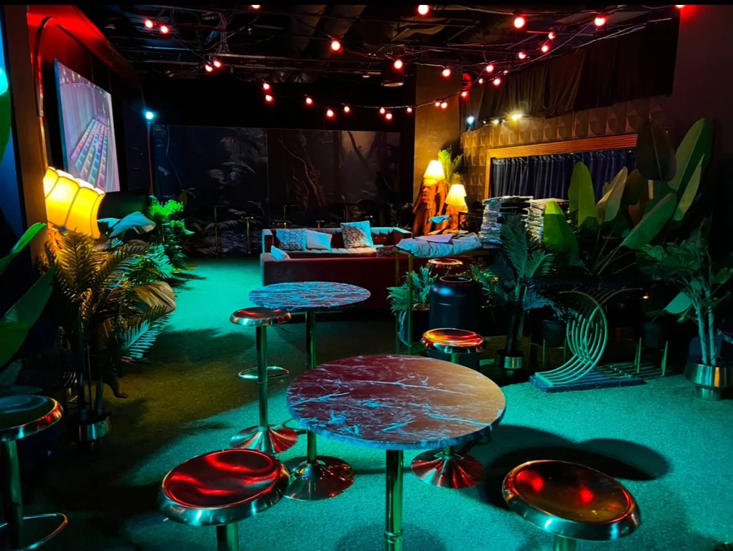 Finished tropical lounge installation with teal flooring, red seating, sculptural plants, and warm string lights